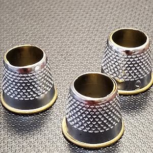 May include: Three silver thimbles with a gold rim. The thimbles are on a textured gray surface.