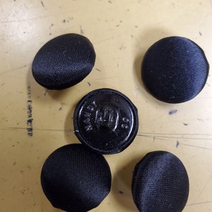 Peut inclure: Cinq boutons noirs, quatre sont ronds et recouverts d'un tissu doux, un est rond et noir avec le texte "HAND MADE" et "30" imprimé dessus.