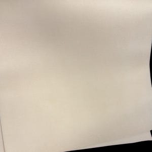 Könnte beinhalten: Ein Blatt cremefarbenes Leder mit leicht strukturierter Oberfläche. Das Leder ist gefaltet, wodurch ein Blick auf die Unterseite freigegeben wird. Der Hintergrund ist schwarz.