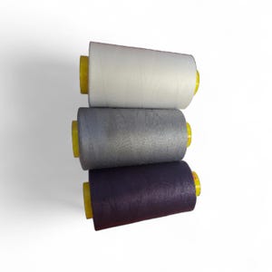 Op de afbeelding: Drie garenspoelen in een verticale stapel. De bovenste spoel is wit, de middelste grijs en de onderste donkerpaars. Elke spoel heeft een gele kern. De achtergrond is wit.