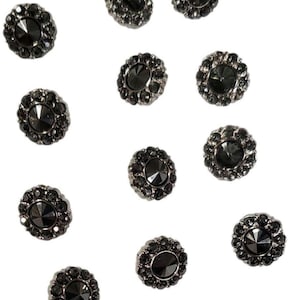 May include: A collection of dark gray, decorative buttons with a floral design. Each button features a central, faceted gem surrounded by smaller, circular embellishments. The buttons are scattered against a white background.