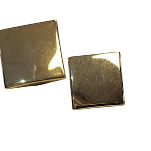 May include: A pair of square, gold-coloured earrings. Each earring has a smooth, reflective surface and slightly rounded edges. The earrings are set against a white background, highlighting their metallic sheen.
