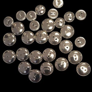 Peut inclure: Un ensemble de boutons argentés à deux trous chacun. Les boutons sont ronds et ont un bord légèrement surélevé. Certains boutons ont un motif décoratif autour du bord.