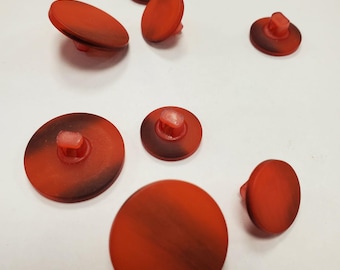 Peut inclure: Un ensemble de six boutons ronds de tailles variées, présentant un motif marbré rouge translucide et brun foncé. Chaque bouton possède une surface lisse et plate ainsi qu'une tige en plastique transparent au dos pour la fixation.