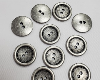 Könnte beinhalten: Eine Sammlung von zehn runden, silberfarbenen Metallknöpfen mit einem abgenutzten Antik-Finish. Jeder Knopf verfügt über zwei mittige Löcher zur Befestigung. Einige Knöpfe haben eine flache Oberfläche, andere einen erhöhten Rand.