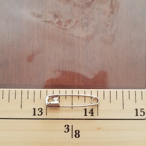 May include: A silver safety pin measuring approximately 1 1/8 inches long, placed on a wooden ruler with markings from 13 to 15.