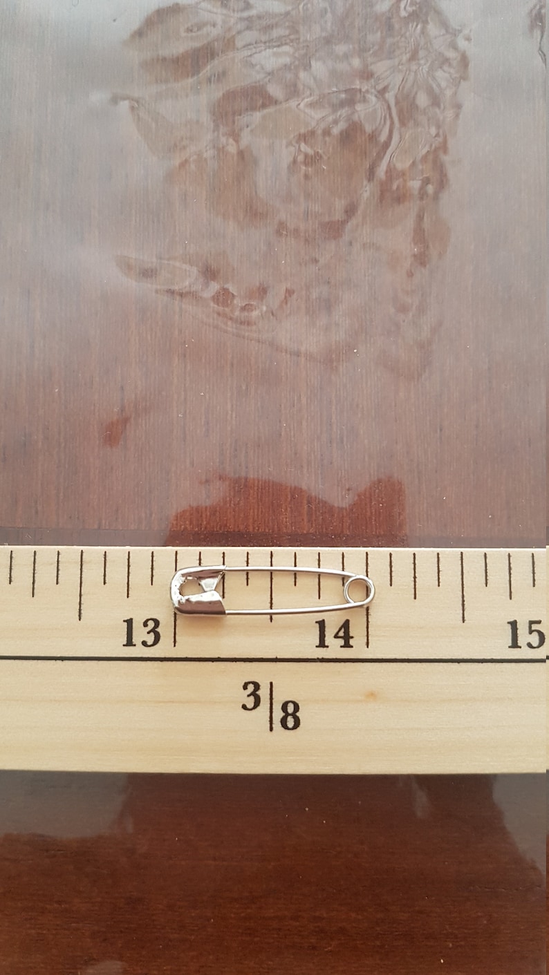 May include: A silver safety pin measuring approximately 1 1/8 inches long, placed on a wooden ruler with markings from 13 to 15.