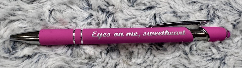 Pu&ograve; includere: Una penna rosa con una clip argentata e una fascia argentata. La penna ha le parole "Eyes on me, sweetheart" incise.
