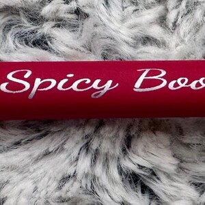 Pu&ograve; includere: Una penna rossa con una clip argentata e una punta argentata. La penna ha le parole "Spicy Book Club" incise sul lato in bianco.