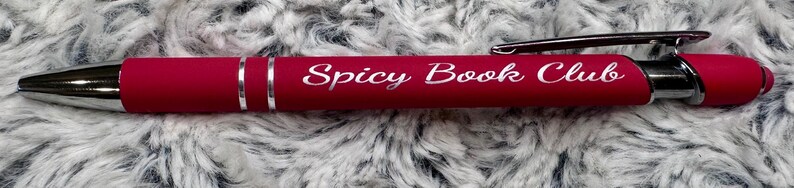Pu&ograve; includere: Una penna rossa con una clip argentata e una punta argentata. La penna ha le parole "Spicy Book Club" incise sul lato in bianco.