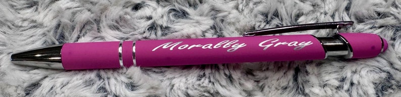 Pu&ograve; includere: Una penna rosa con una clip argentata e una punta argentata. La penna &egrave; incisa con le parole "Morally Gray".