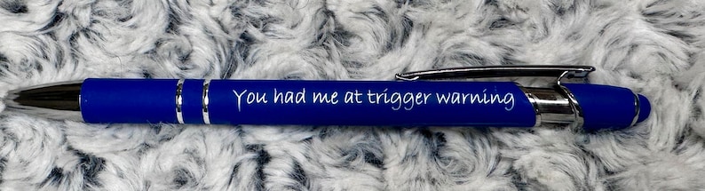 Pu&ograve; includere: Una penna blu con dettagli argentati e l'iscrizione "You had me at trigger warning" sul fusto.