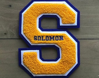 Op de afbeelding: Een gele viltletter 'S' met een witte rand en een donkerblauwe rand. De letter is gestikt met het woord "SOLOMON" in donkerblauw garen.
