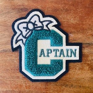 Peut inclure: Écusson vert et blanc avec le mot "CAPTAIN" en lettres blanches. La lettre "C" est en tissu vert texturé. Un nœud blanc est en haut de l'écusson, et l'écusson est bordé de noir.