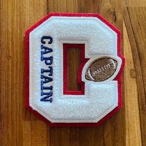 May include: An embroidered patch in the shape of the letter "C", with white, red, and blue detailing. The word "CAPTAIN" is stitched in blue. A brown American football is on the right side.