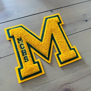 May include: A yellow felt letter M with a green felt outline. The letter has the text "MCHS" embroidered on the left side.