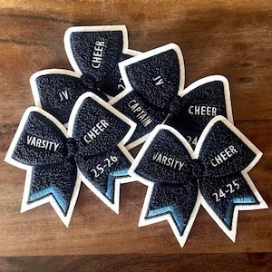 May include: A selection of navy blue cheerleading bows with white edging. The bows feature embroidered text including "CHEER", "VARSITY", "JV", and "CAPTAIN". Some bows have numbers, and a light blue accent.