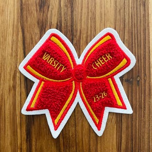May include: A red and yellow Varsity Cheer bow patch with white trim. The patch features the words "VARSITY" and "CHEER" in yellow, with the year "23-26" below. The bow is on a wooden background.