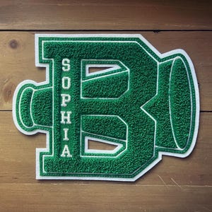 Op de afbeelding: Groen en wit geborduurde patch in de vorm van een gestileerde letter "B" met de naam "Sophia" verticaal. De patch heeft een getextureerde, viltachtige uitstraling en is geplaatst tegen een houten achtergrond.