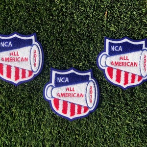 May include: Three embroidered patches with a shield shape, featuring a white megaphone and the text "NCA ALL AMERICAN" in red and white. The patches have the years 2018, 2019, and 2022. The patches are on a green background.
