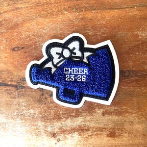 May include: A blue and white embroidered patch in the shape of a megaphone. The patch has the word "CHEER" and the numbers "23-26" embroidered in white. The patch is outlined in black and white.