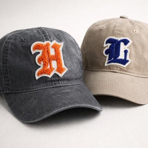 May include: Two baseball caps. One is charcoal grey with an orange "H" patch, the other is tan with a blue "L" patch. Both caps have a curved brim and adjustable strap.