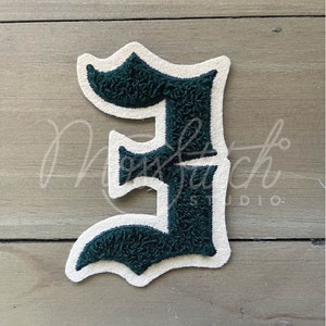 Pode incluir: Um patch de letra "E" verde escuro com uma borda branca. A letra tem uma aparência texturizada e difusa. O patch está em uma superfície de grão de madeira clara. O logotipo "Moss Stitch Studio" está no canto inferior direito.