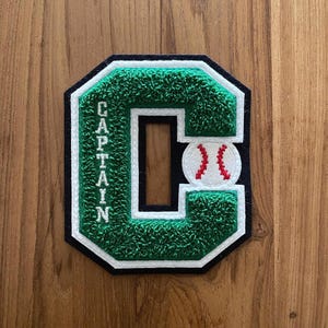 Op de afbeelding: Een groen, wit en zwart geborduurd embleem in de vorm van de letter "C". Het woord "CAPTAIN" is verticaal in het wit geborduurd. Een honkbal met rode stiksels staat rechts.