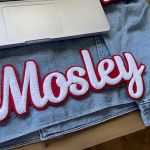 Puede incluir: Una chaqueta vaquera con un parche bordado grande que dice "Mosley" en letras blancas con un borde rojo. Un ordenador portátil plateado descansa sobre la chaqueta, que está colocada sobre una superficie de madera.