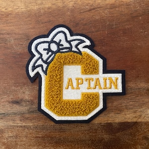 Könnte beinhalten: Ein gold-weiß gestickter Aufnäher mit dem Wort „CAPTAIN“ im College-Schriftstil. Der Aufnäher hat einen marineblauen Rand und ein weißes Schleifendesign oben. Der Aufnäher ist wahrscheinlich für eine Jacke oder eine Tasche.