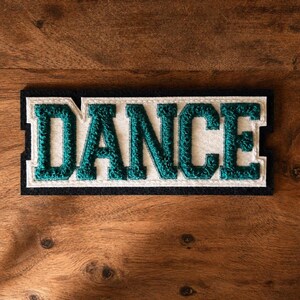 May include: A rectangular patch featuring the word DANCE in textured teal chenille lettering. The letters are outlined in white felt, which is set against a solid black rectangular background, creating a layered varsity-style appearance.