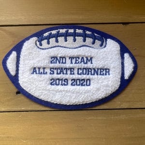 May include: A blue and white embroidered patch in the shape of a football. The patch reads "2ND TEAM ALL STATE CORNER 2019 2020" in blue thread. The patch is on a wooden surface.