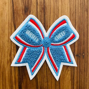 May include: A blue, red, and white embroidered cheerleading bow patch. The bow features the words "VARSITY CHEER" in white lettering. The patch has a textured, fuzzy appearance and is set against a wooden background.