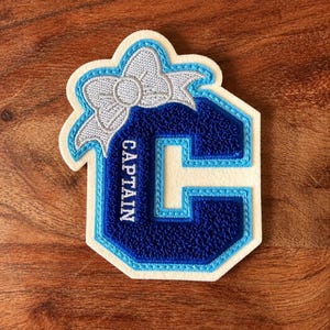 May include: A patch featuring a large, dark blue letter C with a light blue border on a cream-coloured felt base. The letter is embroidered with the word CAPTAIN in white thread. A silver-grey embroidered bow sits atop the letter C.
