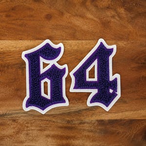 May include: A pair of embroidered patches featuring the number "64" in a gothic font. The numbers are purple with a textured appearance and a white border. The patches are set against a wooden background.