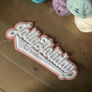 Può includere: Un cerotto in feltro rosa e bianco con il testo "Fabulous" in un font corsivo. Il cerotto ha la forma di una palla da baseball.