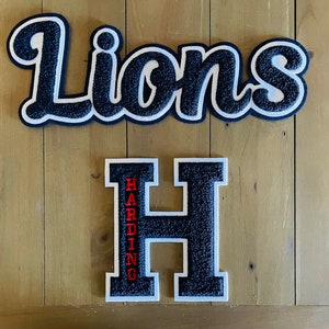 Op de afbeelding: Twee patches in varsity-stijl. De bovenste patch leest "Lions" in een marineblauw, wit en zwart ontwerp. Hieronder staat een zwarte "H" patch met witte rand en het woord "Harding" in rood.