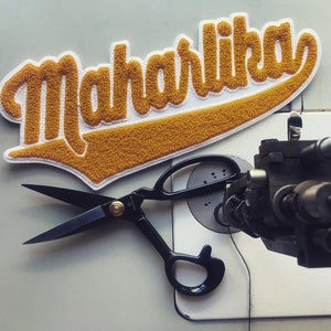 May include: A gold and white felt patch with the word "Maharlika" stitched on it. A black pair of fabric shears are lying on a white surface next to the patch.