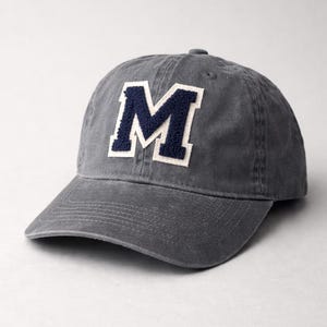 May include: Gray baseball cap with a curved brim. The cap features a large, navy blue letter "M" with a white border on the front. The hat is made of a soft material and has a button on top.