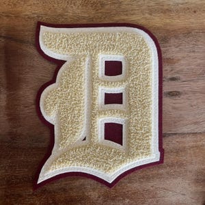 May include: A varsity letter patch in the shape of the letter "D". The letter is beige with a textured, fuzzy appearance, outlined in white and maroon. The patch has a maroon border and is likely for a jacket or apparel.