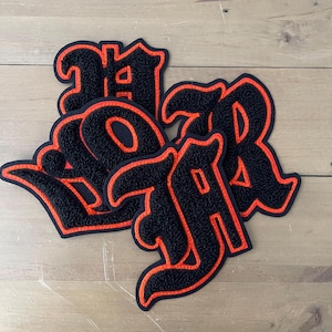 Könnte beinhalten: Vier schwarze Filzpatches mit orangefarbener Steppnaht, jeder mit einem anderen Buchstaben in gotischer Schrift. Die Buchstaben ergeben "WAR".