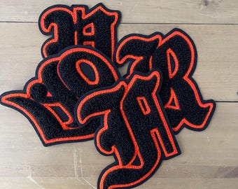 May include: Four black felt patches with orange stitching, each featuring a different letter in a gothic font. The letters spell out "WAR".
