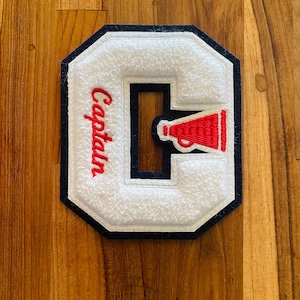Op de afbeelding: Een witte vilten letter "C" patch met een rode megafoon en het woord "Captain" in rood script. De patch heeft een marineblauwe rand en is geplaatst tegen een houten achtergrond.