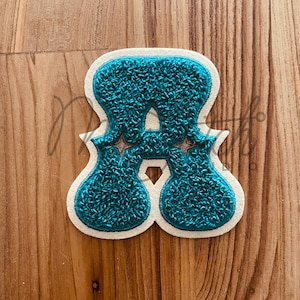 Könnte beinhalten: Türkisfarbener Chenille-Buchstaben-A-Aufnäher mit cremefarbenem Rand und Sternakzenten. Der Aufnäher hat ein strukturiertes, flauschiges Aussehen und eignet sich für Kleidung oder Bastelarbeiten.