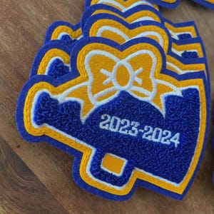 May include: A collection of blue, gold, and white embroidered patches. Each patch features a bow design in gold and white, with the text "2023-2024" in white. The patches have a unique shape with a blue border and gold accents.