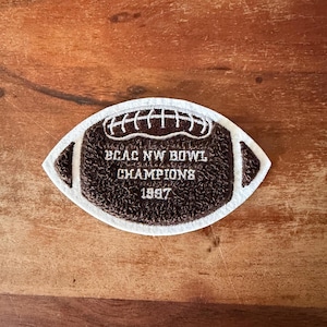 May include: A brown and white football-shaped patch with the text "BCAC NW BOWL CHAMPIONS 1997". The patch has a textured appearance and is likely an embroidered item.