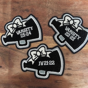 May include: Three black and white megaphone patches with text. Each patch has a bow at the top and a silver outline. The text on the patches reads "VARSITY 23-24", "VARSITY 22-23", and "JV 21-22".