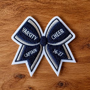 May include: Navy blue and white embroidered cheerleading bow with the words "VARSITY CAPTAIN" and "CHEER 26-27". The bow is on a wooden surface.