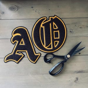 Puede incluir: Dos parches de fieltro negro con costuras amarillas, uno con la letra "A" y el otro con la letra "O". Los parches están sobre una superficie de madera con un par de tijeras negras.