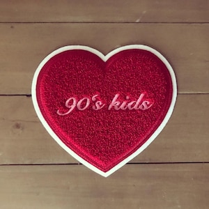 Może przedstawiać: Czerwona łatka w kształcie serca z białym obramowaniem i napisem "90's kids" w różowych literach.